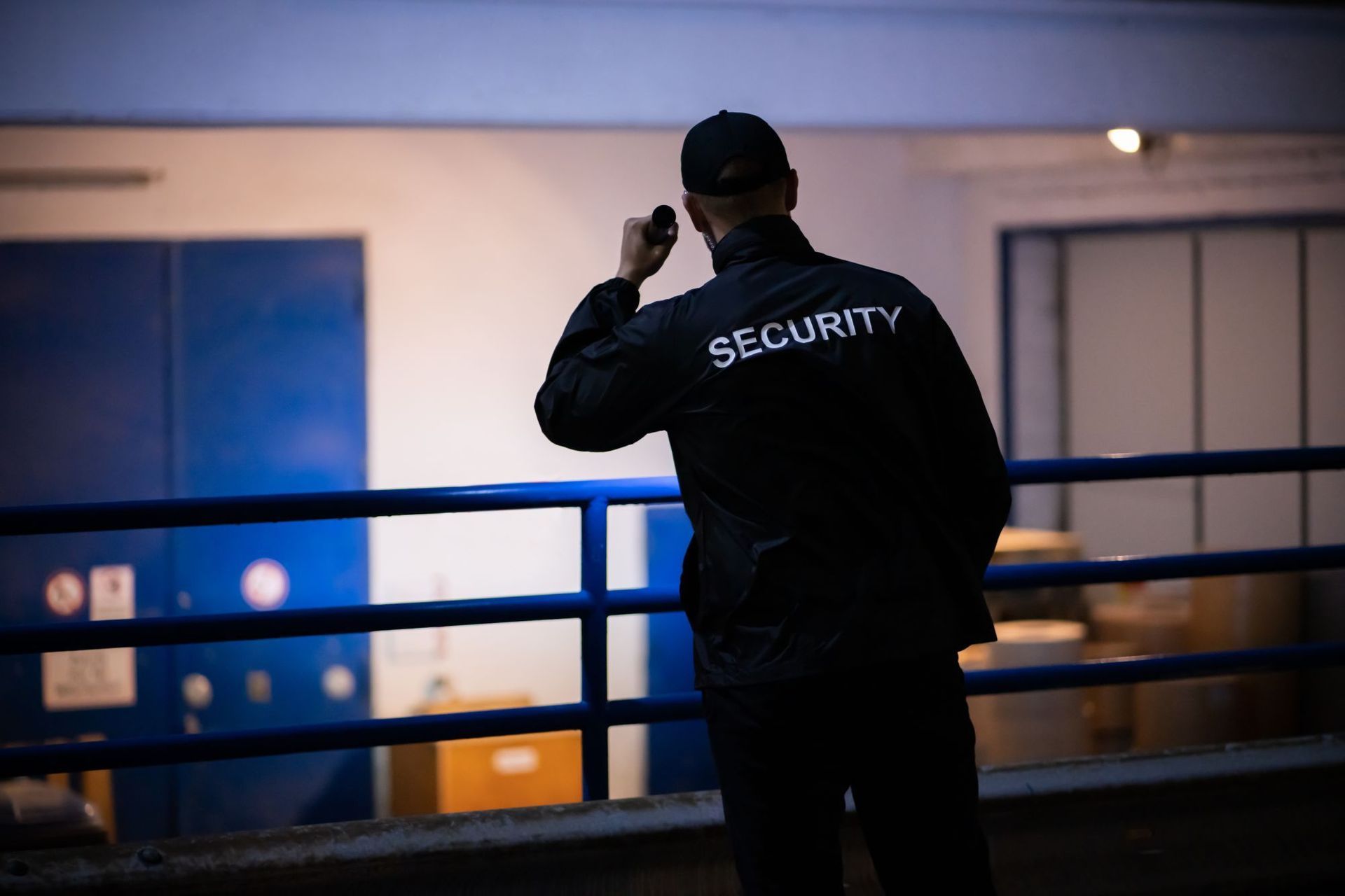 Security guard in dark uniform holding a radio, looking to the side near a blue and white building.