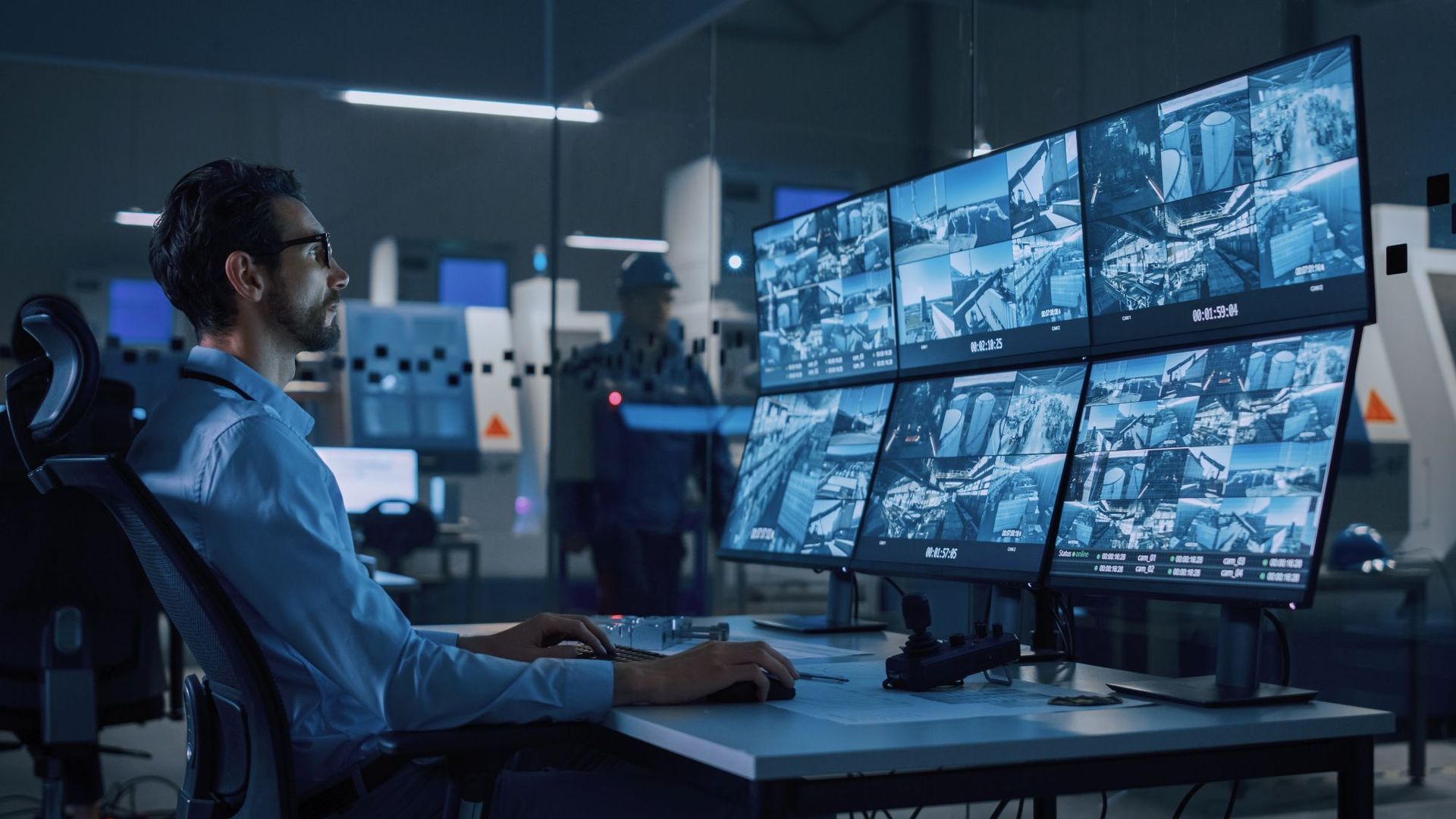 Man monitors security cameras at a control station. Multiple screens show camera views. Dark, blue-lit environment.