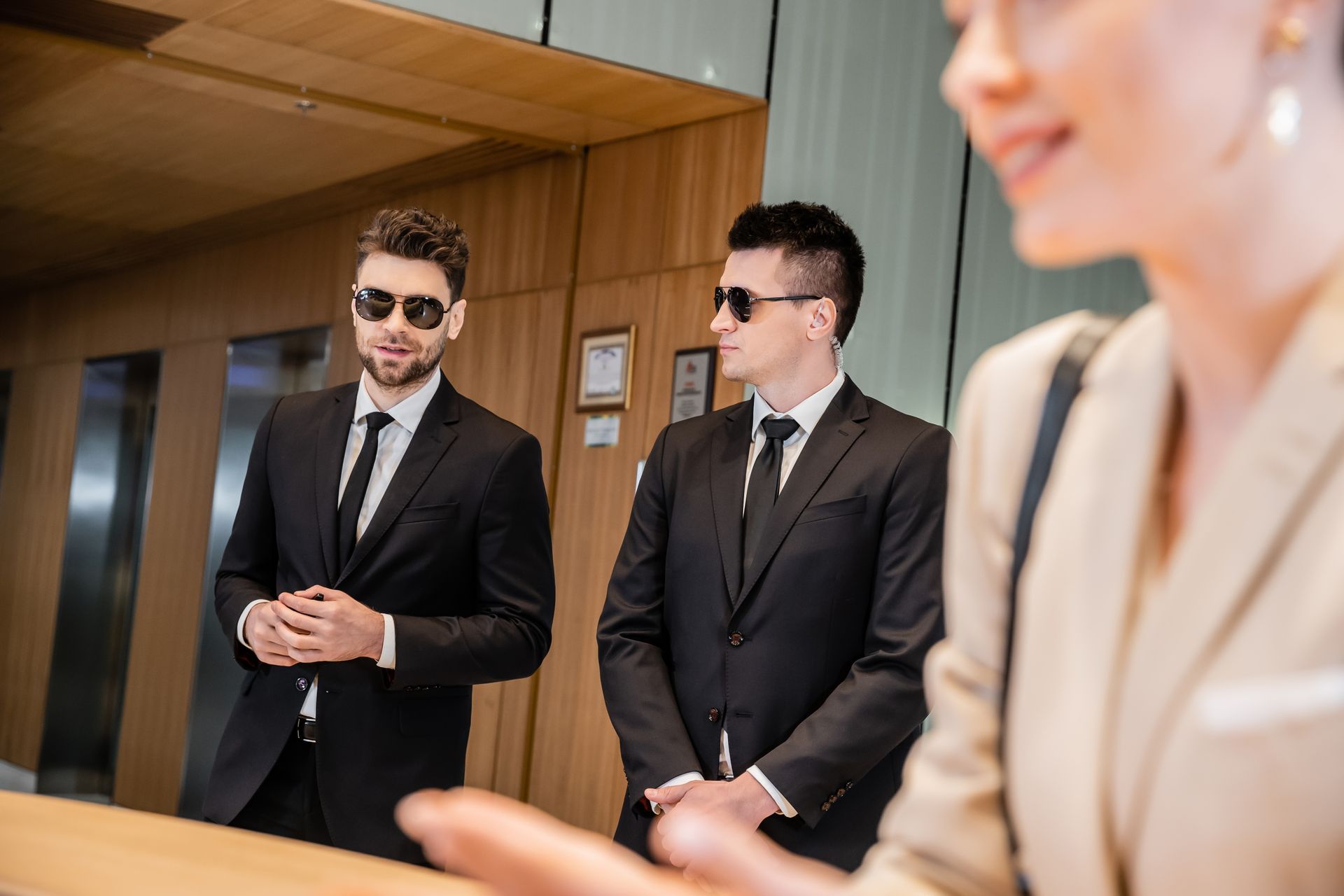 Two suited men with sunglasses stand near an elevator, possibly security, with a woman at a reception desk.