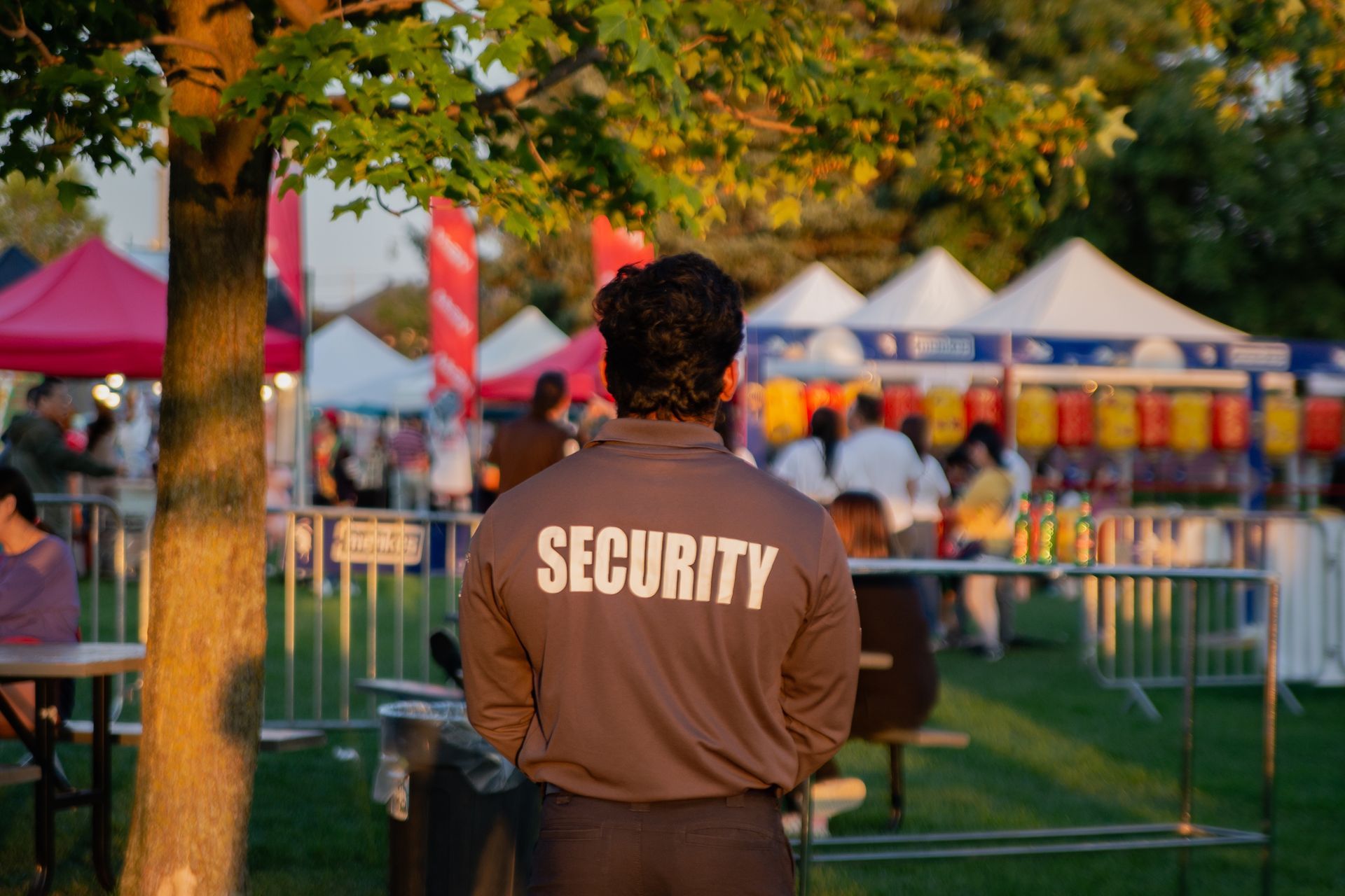 Security guard at an outdoor event, back to the viewer, watching crowd near tents and barriers.