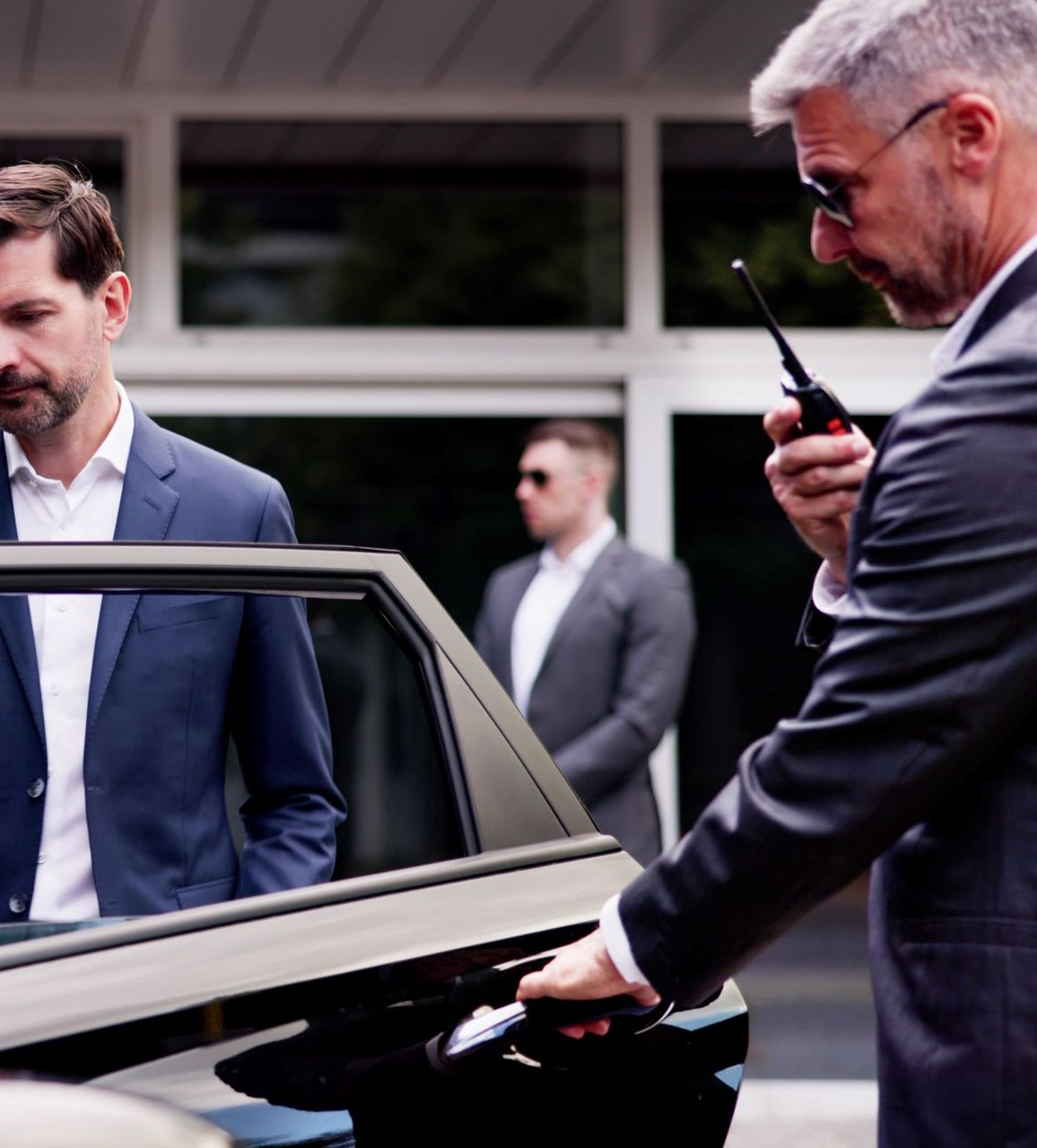 Man in suit about to enter car, guarded by bodyguards with walkie-talkie. Modern building in background.