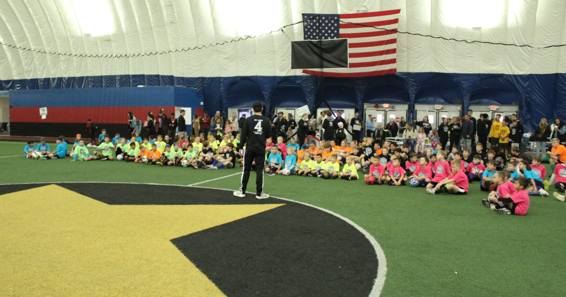 Nick Desrocher standing before a large group of kids in bright jerseys inside Starhill Family Athletic Center; American flag overhead.