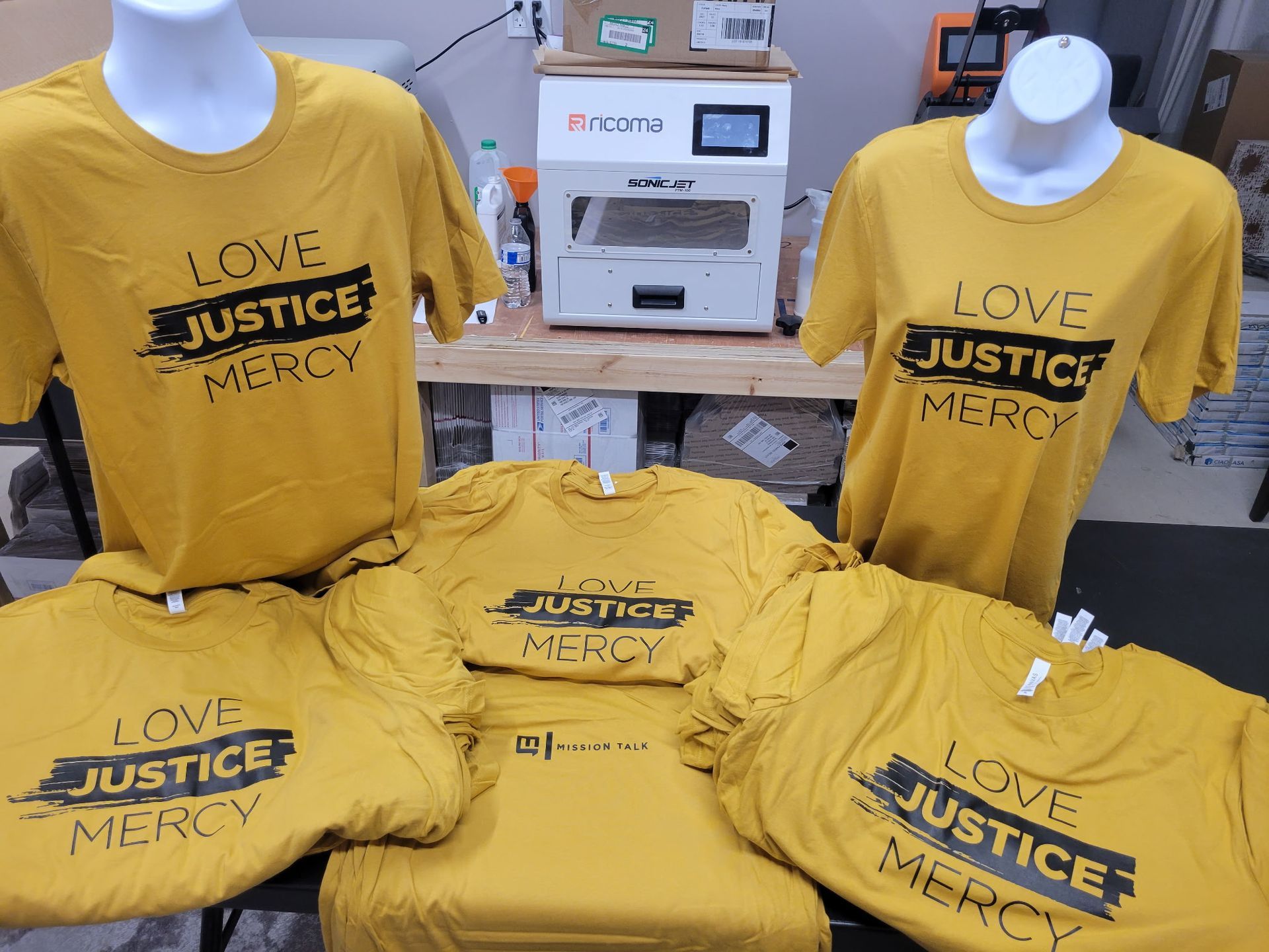 A bunch of yellow t-shirts with the words `` love justice mercy '' on them are sitting on a table.