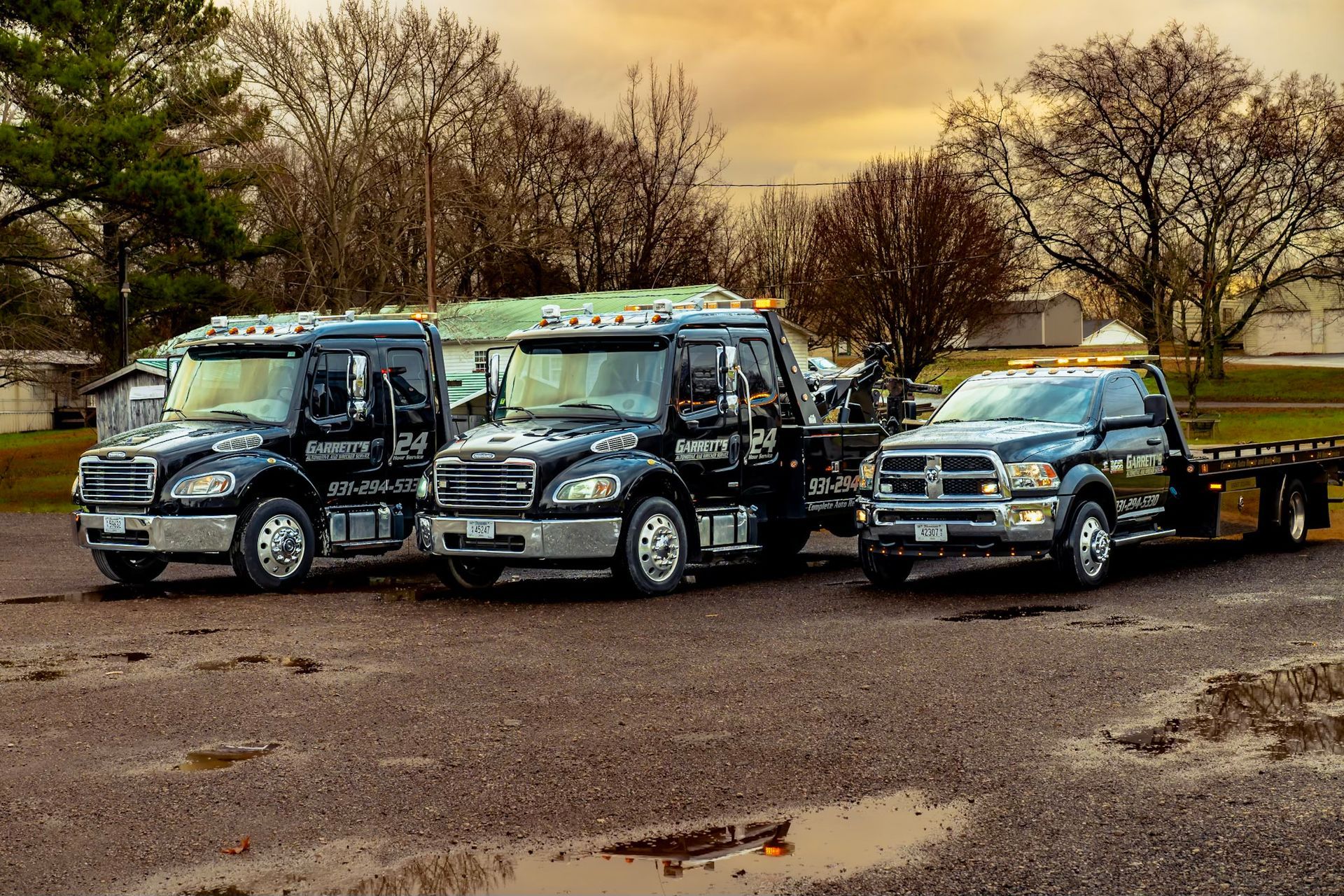 Three black tow trucks parked on a gravel lot under a cloudy sky.