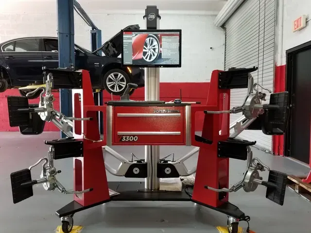 Wheel alignment machine in auto repair shop; car on lift in background. Red and gray machine, monitor displays wheel image.