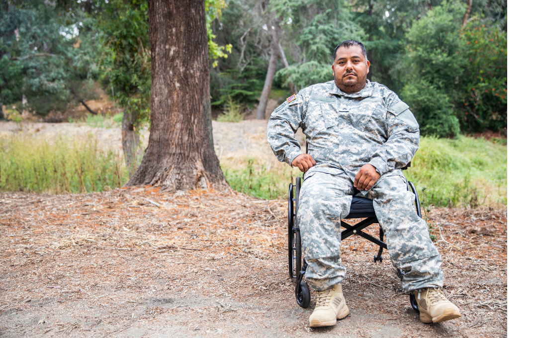 Soldier in camouflage uniform sits in a wheelchair outdoors near a tree.