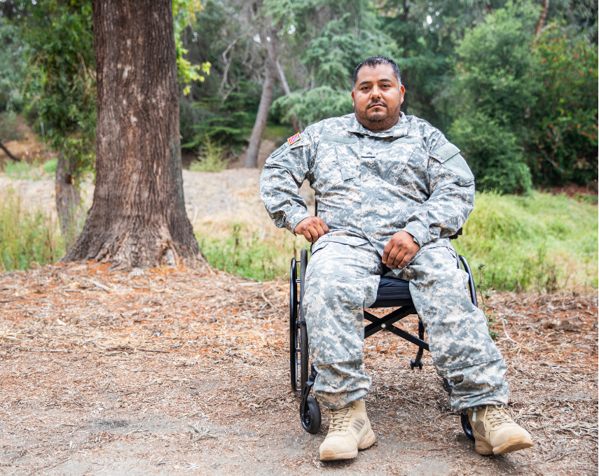 Man in military uniform sits in a wheelchair on a dirt path, near a tree, in a wooded area.
