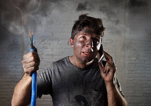 Man covered in soot holding smoking electrical wires, talking on phone.