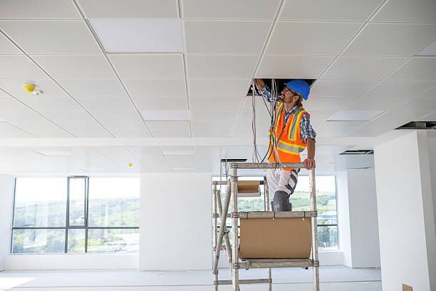Construction worker on a ladder, installing wires in a suspended ceiling. The worker wears a hard hat and safety vest.