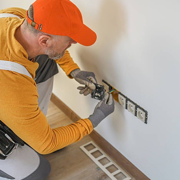 Man in orange hat and gloves installing electrical outlet on a white wall.