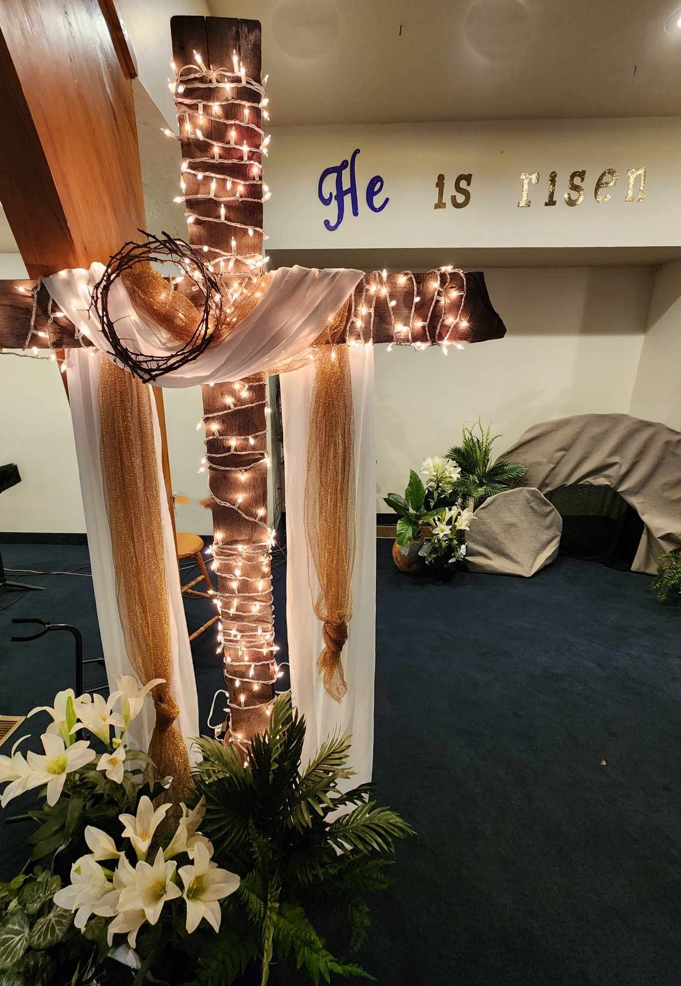 A cross decorated with lights and flowers in a room with the words he is risen on the wall.
