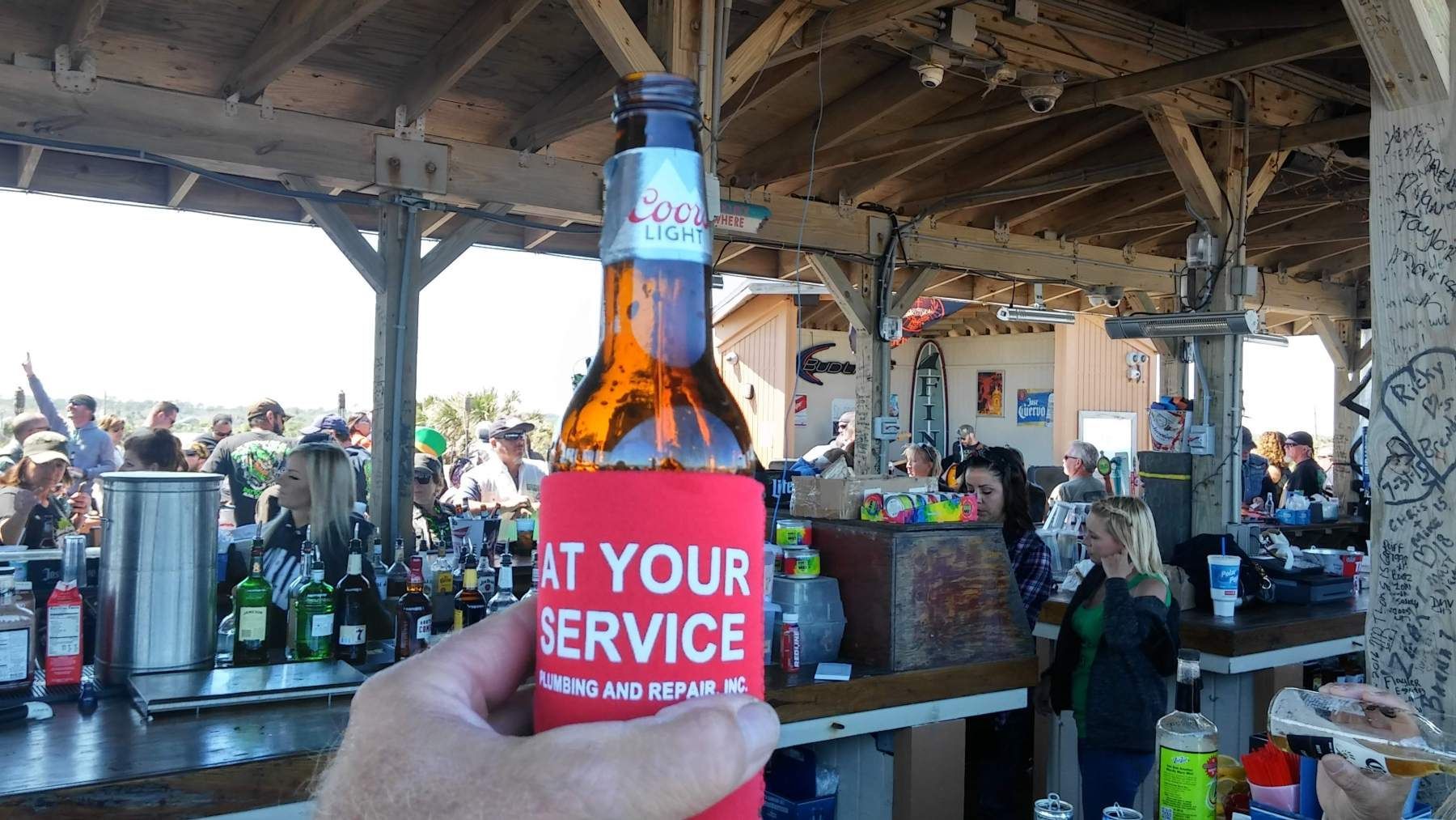 A person is holding a bottle of beer in front of a bar that says at your service