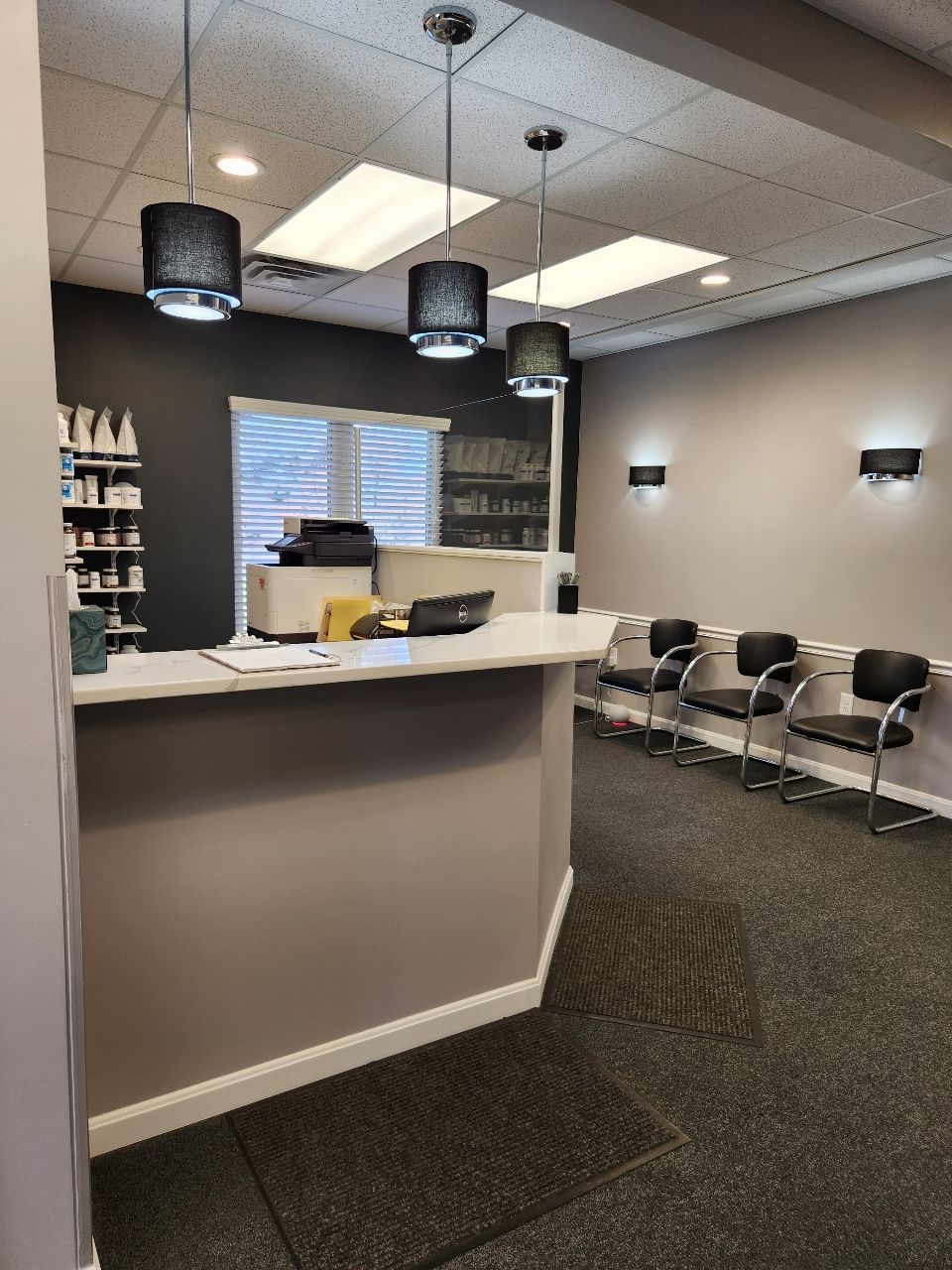 Reception area with desk, chairs, and lighting. Gray walls, dark carpet, and waiting area.