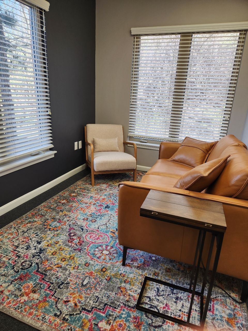 Cozy office corner: tan leather couch, beige armchair, colorful rug, and two windows with blinds.