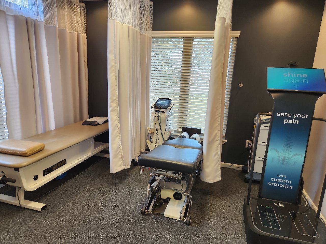 Chiropractic exam room with adjustable tables, dark walls, and a Know Your Pain machine.