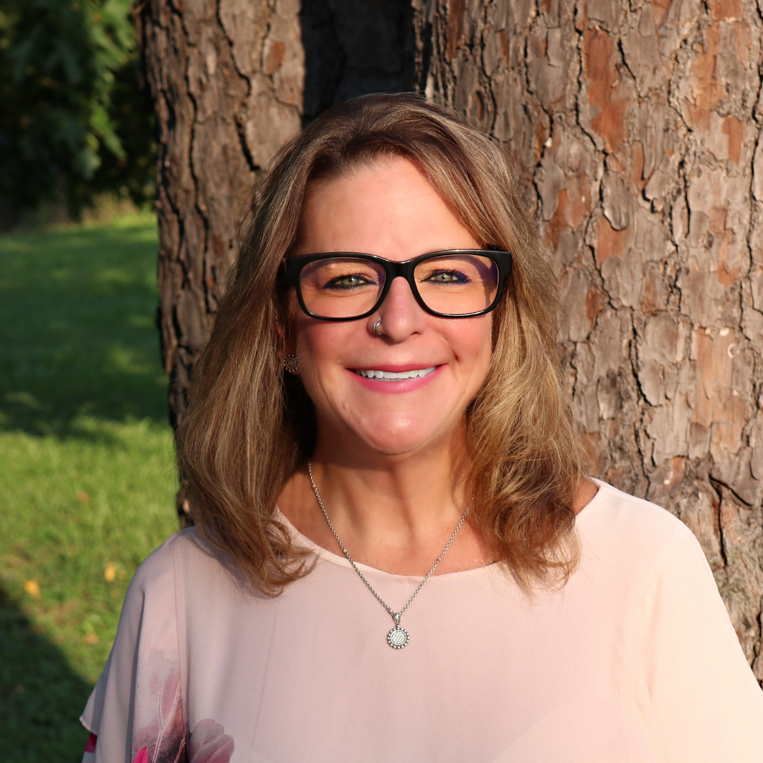 Woman with glasses smiles in front of a tree; wearing a light pink shirt and necklace.