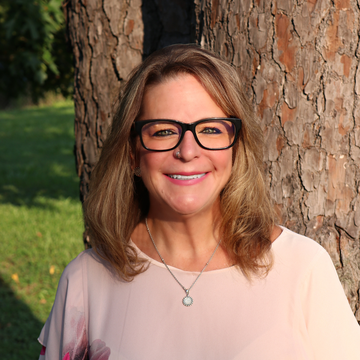 Woman with glasses smiles in front of a tree; wearing a light pink shirt and necklace.
