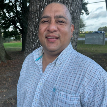 Man with a shaved head, smiling, outdoors near a tree, wearing a button-down shirt.