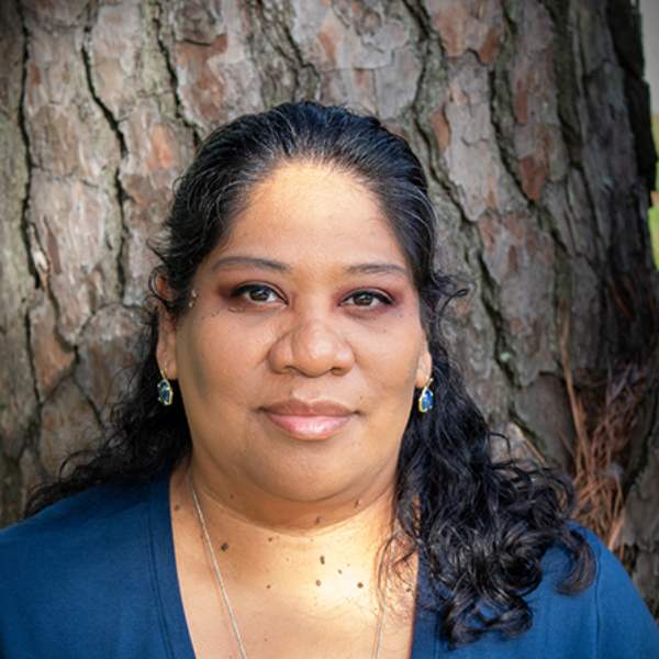 Woman with dark hair, blue top, brown eyes, and small earrings, against a tree trunk.