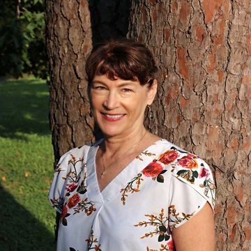 Woman with short brown hair, smiling, in floral blouse, standing next to a tree.