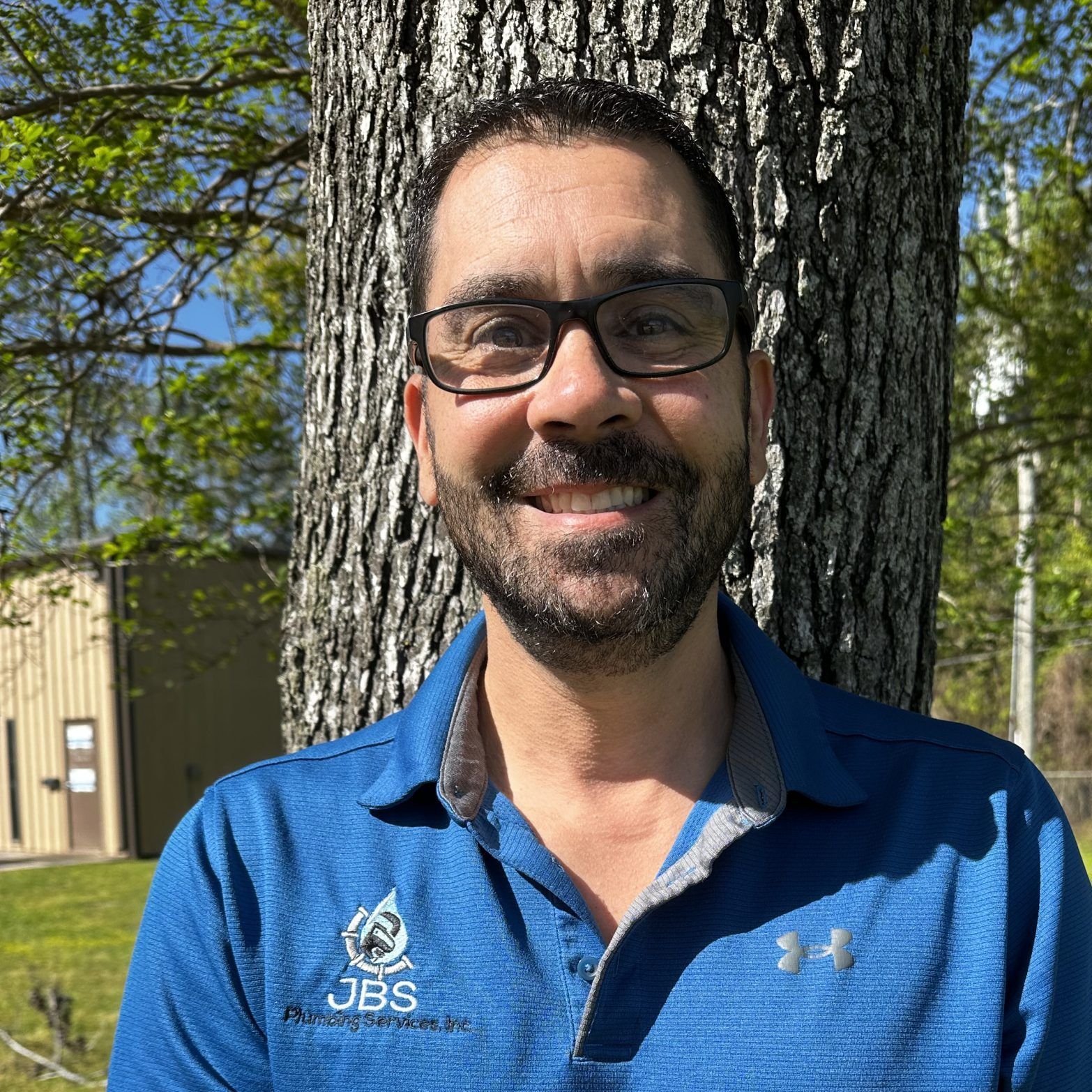 Man with glasses smiles, stands near a tree outdoors, wearing a blue polo shirt.