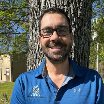 Man with glasses smiles, stands near a tree outdoors, wearing a blue polo shirt.