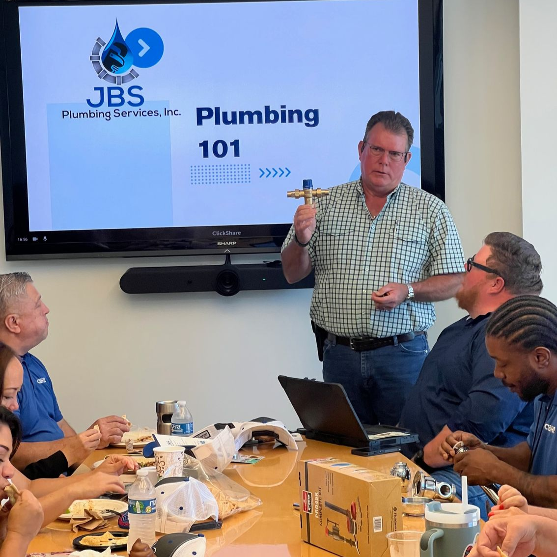 Man teaching a plumbing class, holding a fitting. People seated at table, watching.