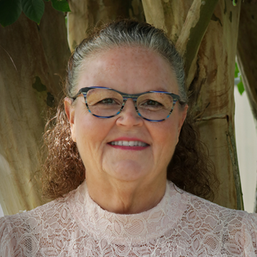 Woman with glasses, wearing a pink lace top, smiles at camera, outdoors with tree in background.