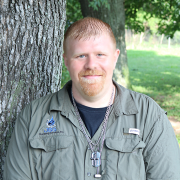 Man with ginger hair and beard, wearing green shirt, leaning against a tree, smiling outdoors.