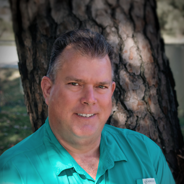 Man in green shirt smiling in front of a tree.