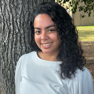 Woman with curly black hair smiles, leaning against a tree in a park; wearing a white top.