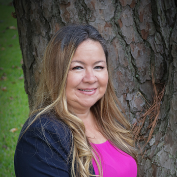 Woman with long brown hair, pink top, and blue blazer smiles, leaning against a tree.