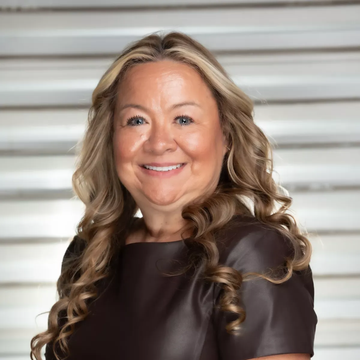 Woman with glasses smiles in front of a tree; wearing a light pink shirt and necklace.