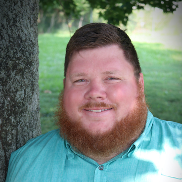 A man with a red beard and a turquoise shirt smiles, leaning against a tree outside.