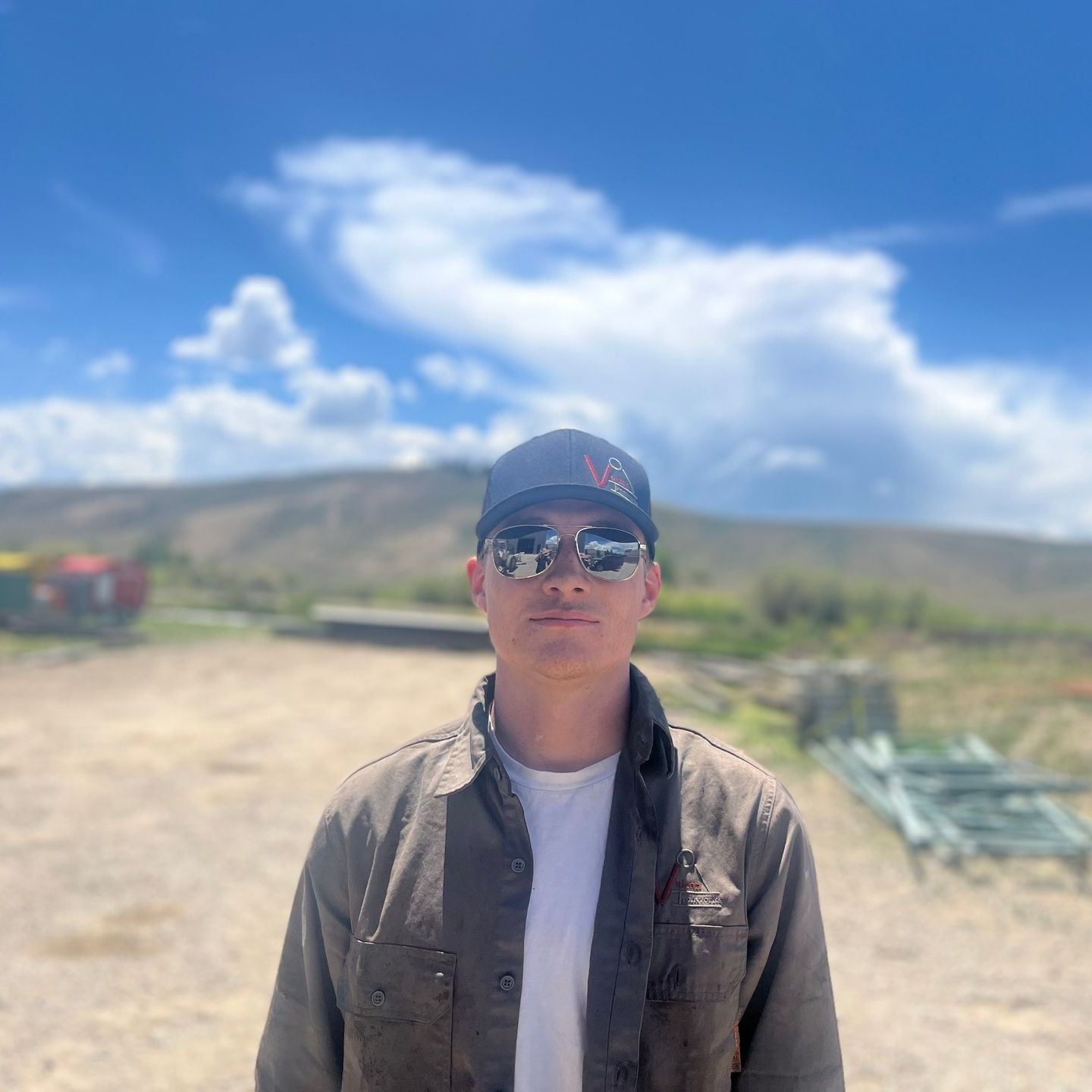 A man wearing a hat and sunglasses is standing in a dirt field.