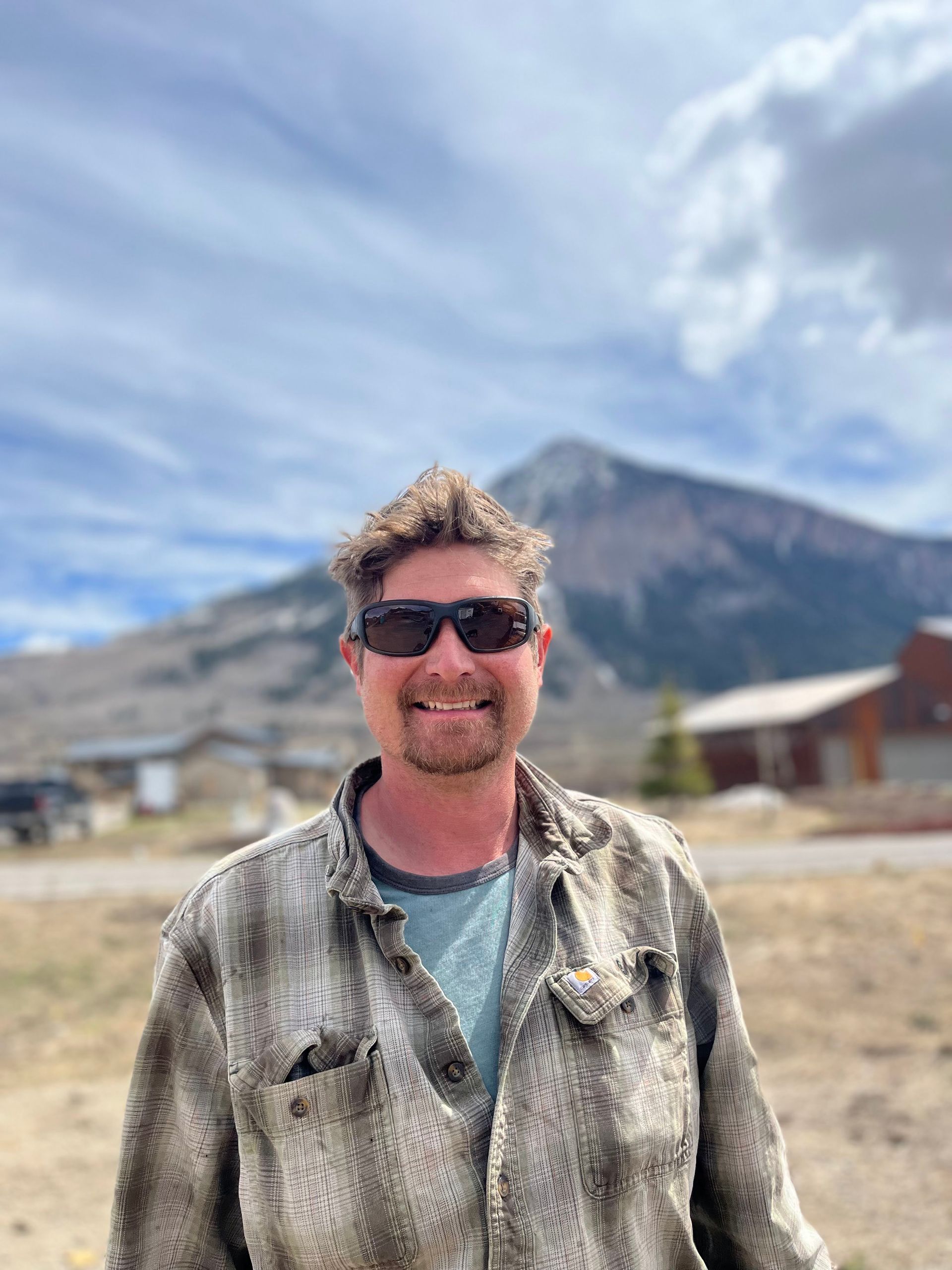 A man wearing sunglasses and a plaid shirt is standing in front of a mountain.