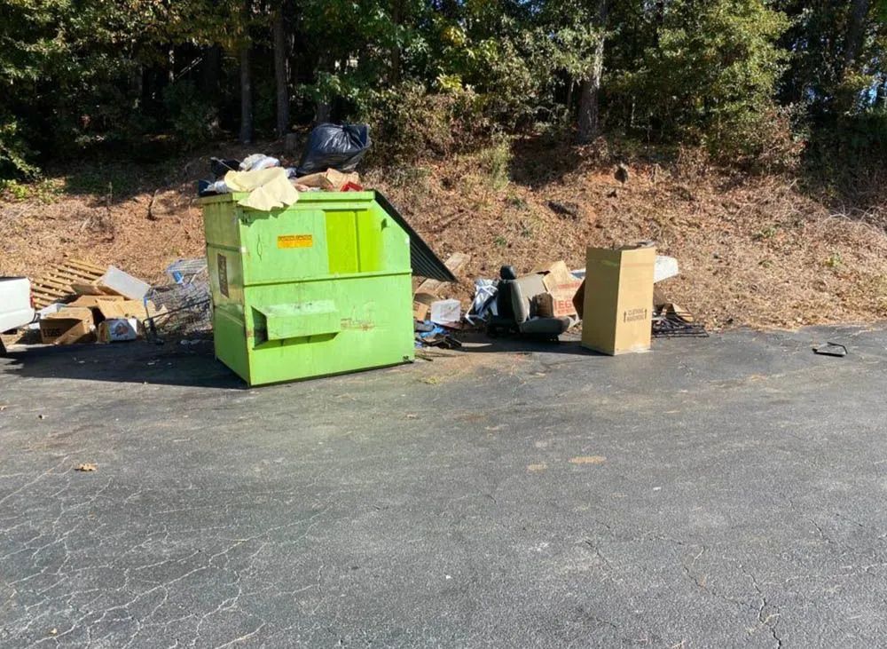 A green dumpster is sitting on the side of the road next to a pile of trash.
