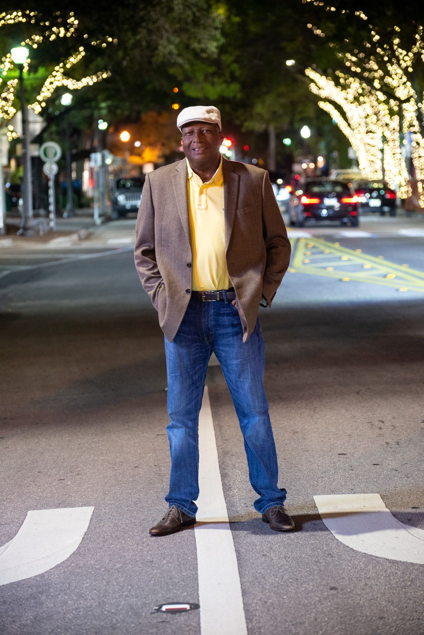 Man in a blazer and jeans stands in a crosswalk at night, city lights in background.