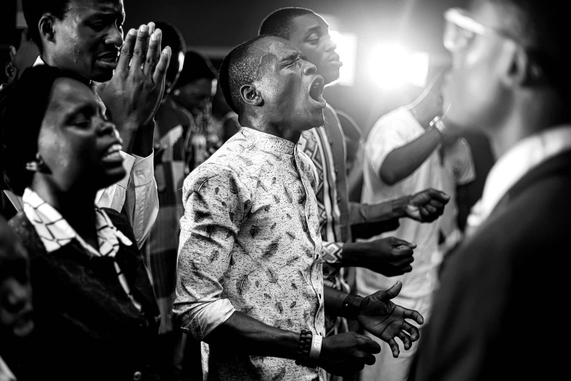 People singing and worshiping in a dimly lit space. Man in patterned shirt shouts; others raise hands.