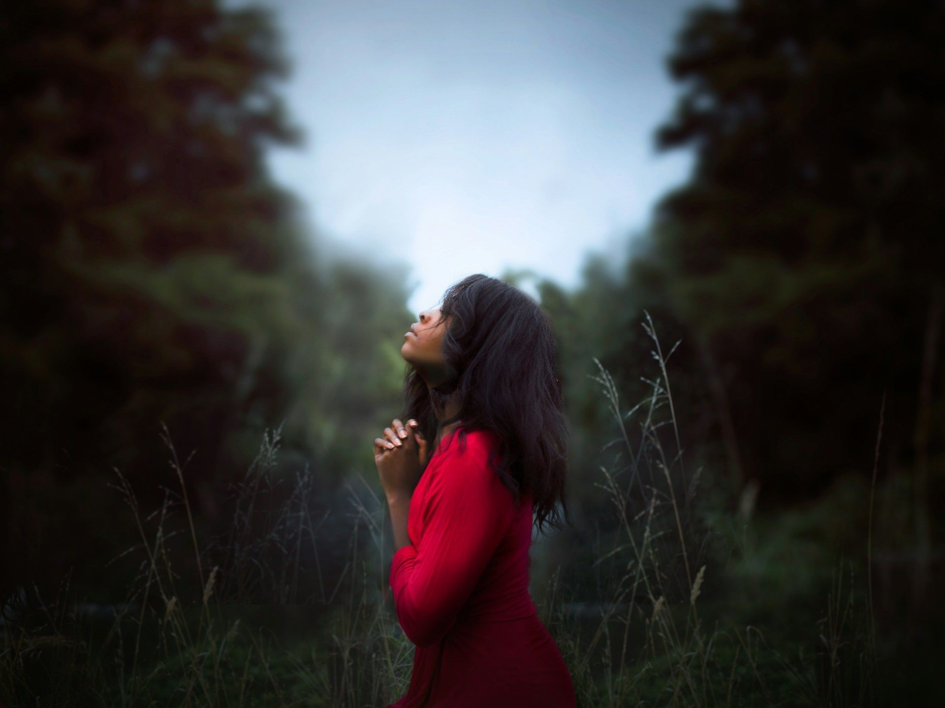 Woman in red dress with hands clasped, looking upward in a forest.