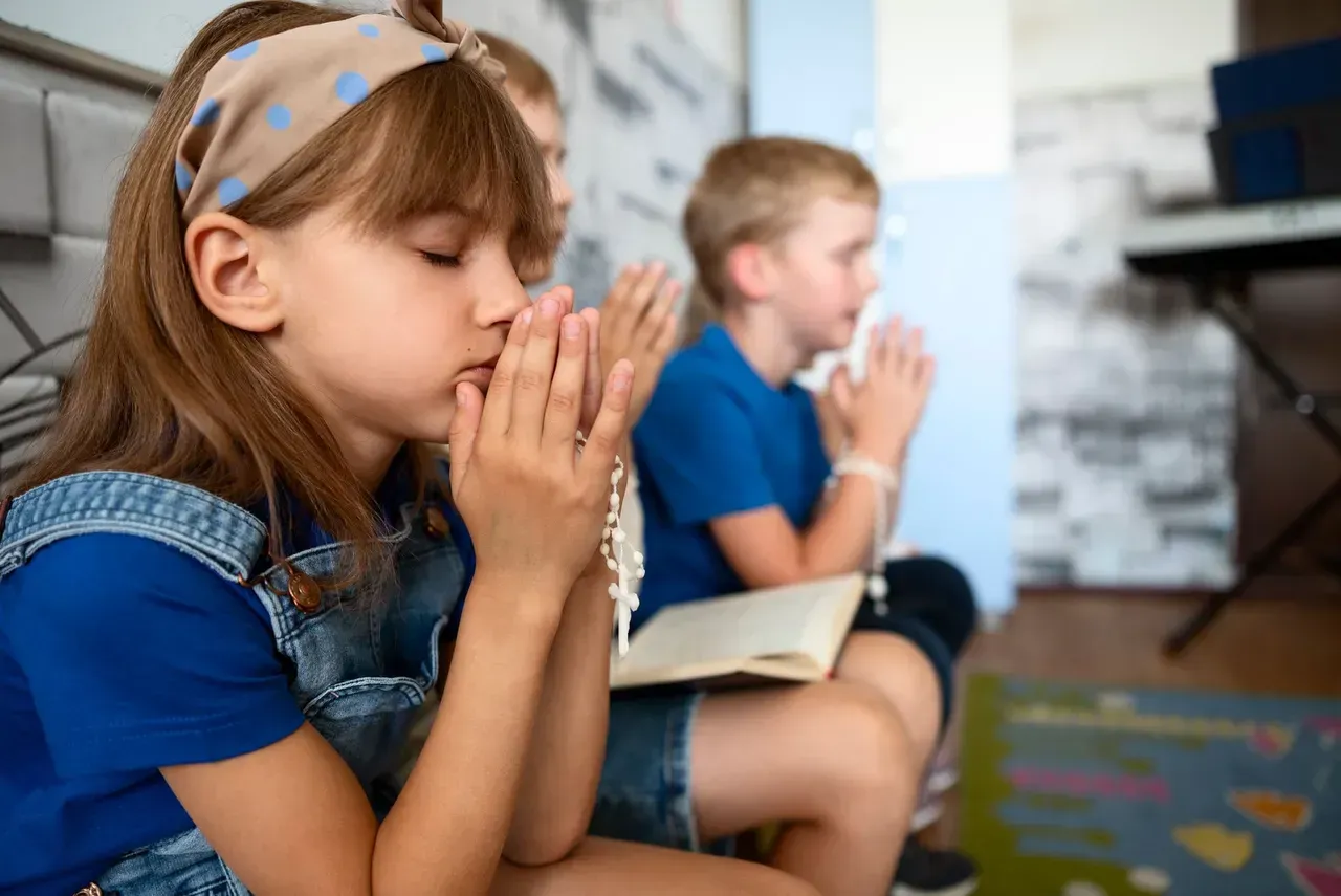 Children with hands clasped in prayer, eyes closed, sitting in a room.