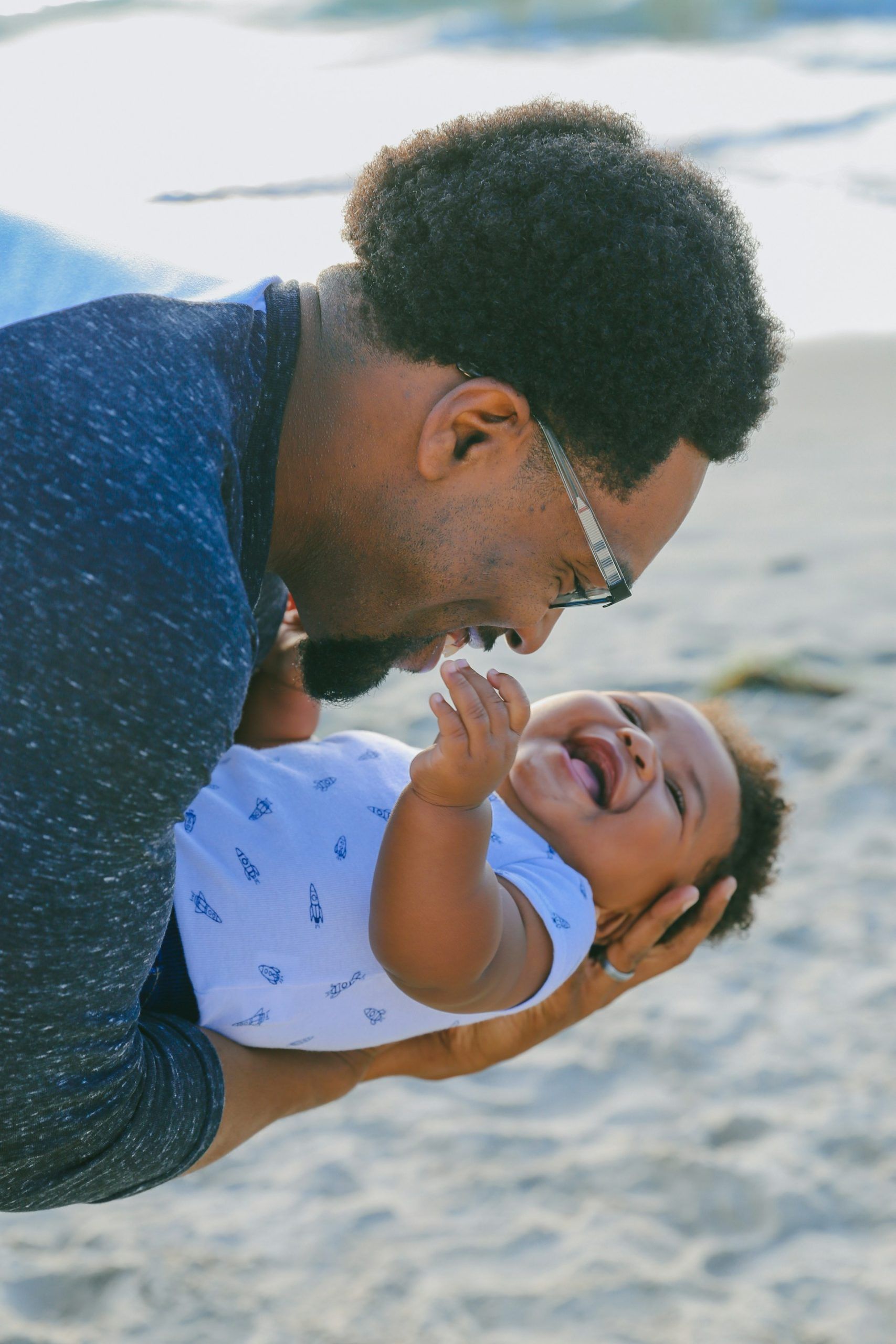 Man holding and playfully smiling at laughing baby on a beach.