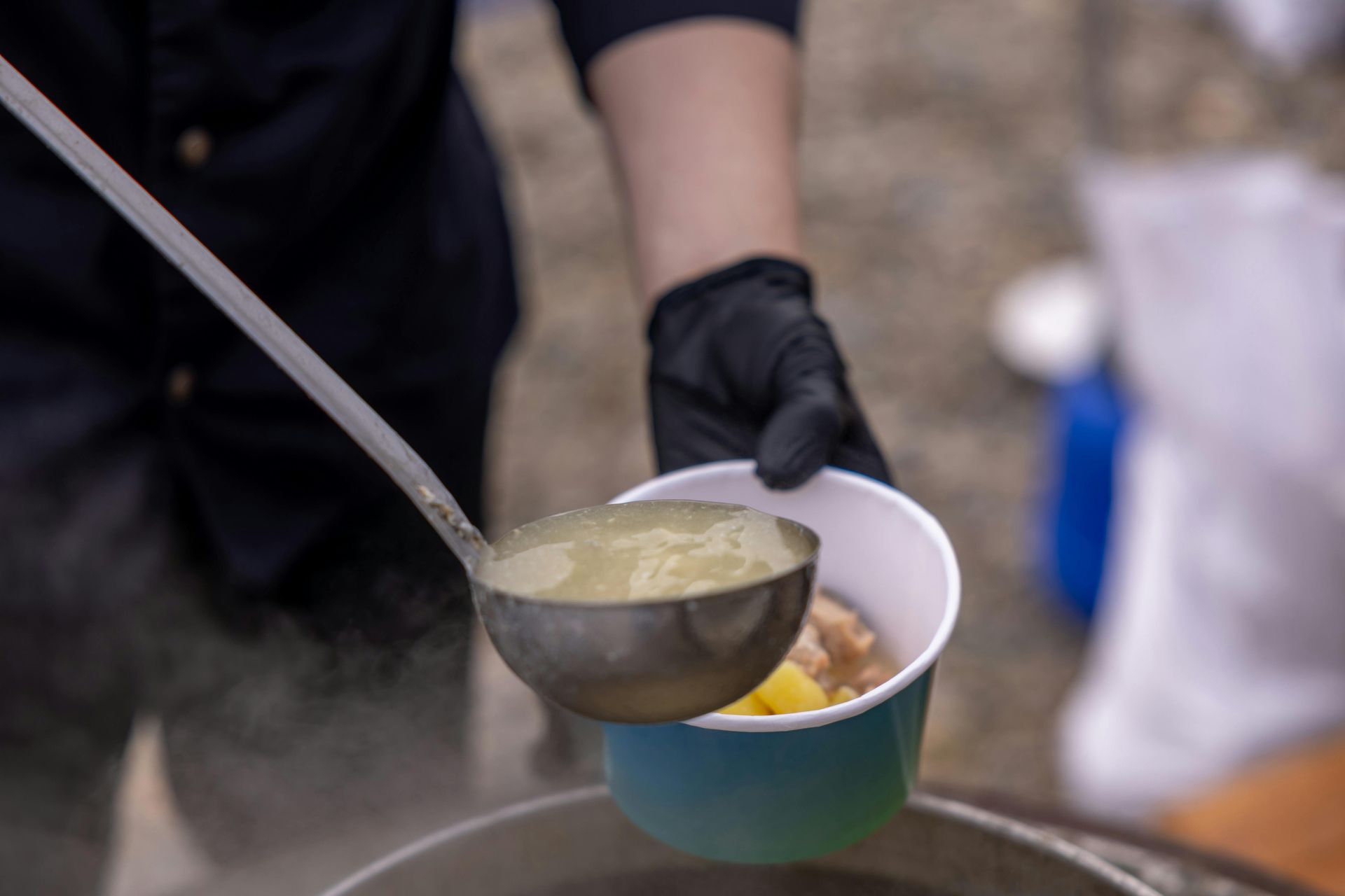 Person in black gloves ladling soup from a pot into a paper bowl.