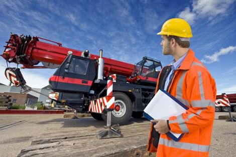 A man wearing a hard hat is holding a clipboard in front of a crane.