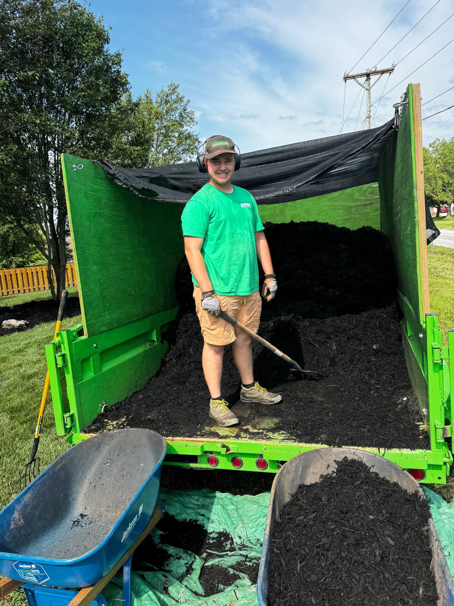 A man in a green shirt is standing in the back of a green dumpster holding a shovel.