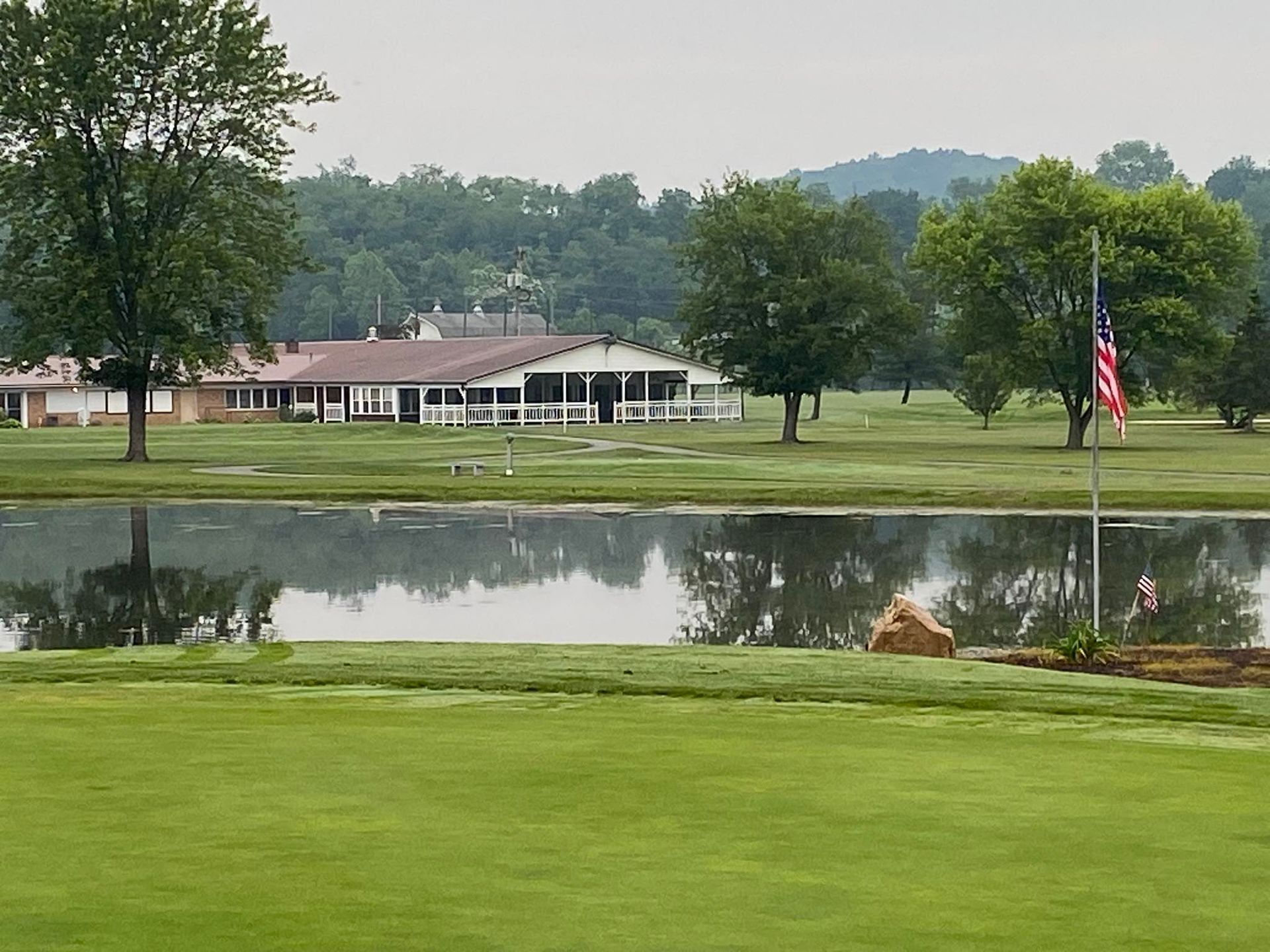 A green golf course with a pond in front of a white building with a porch. An American flag waves.