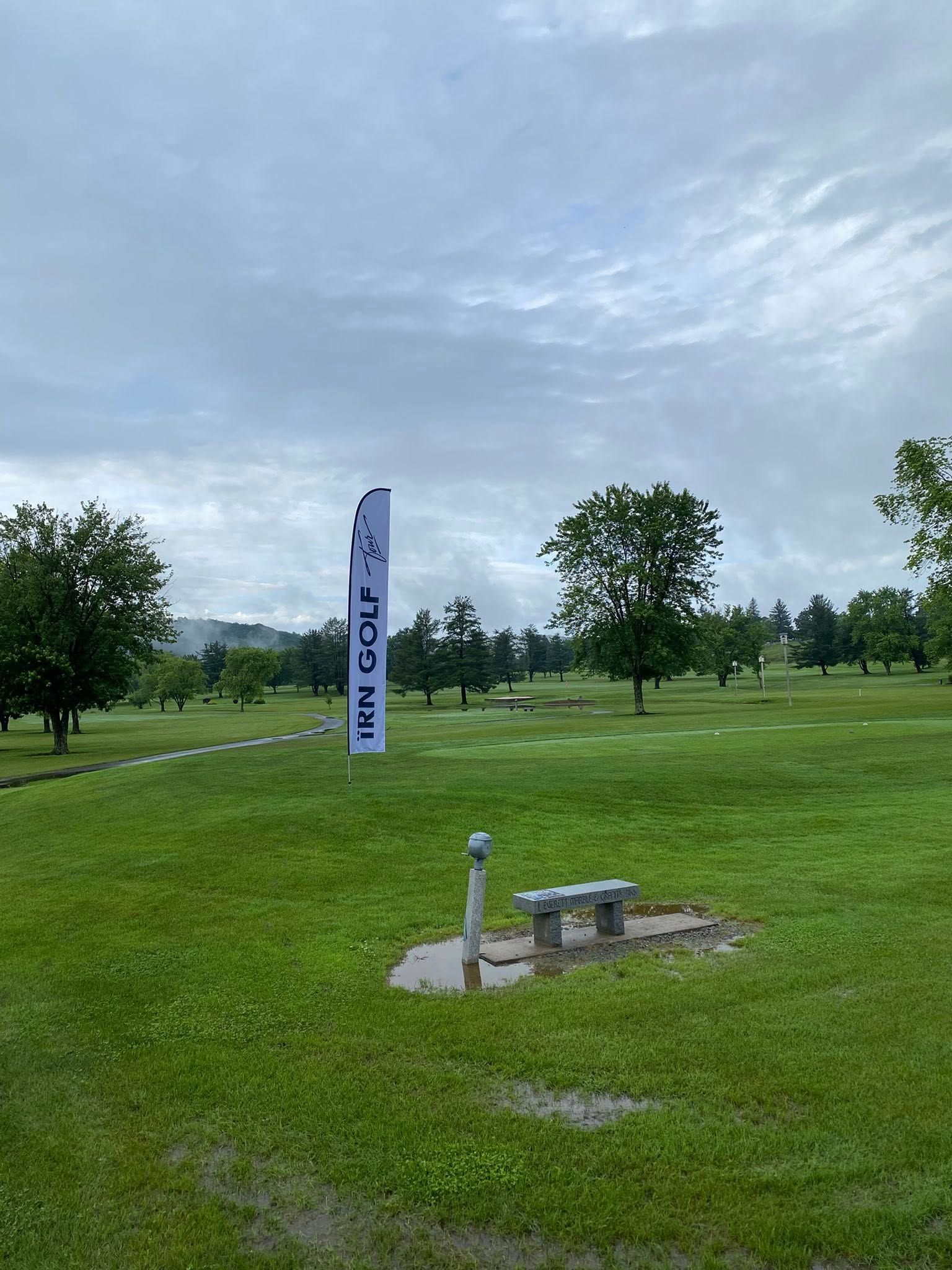 Golf course on a cloudy day. A bench and flag are in the foreground, with trees and the course on the background.