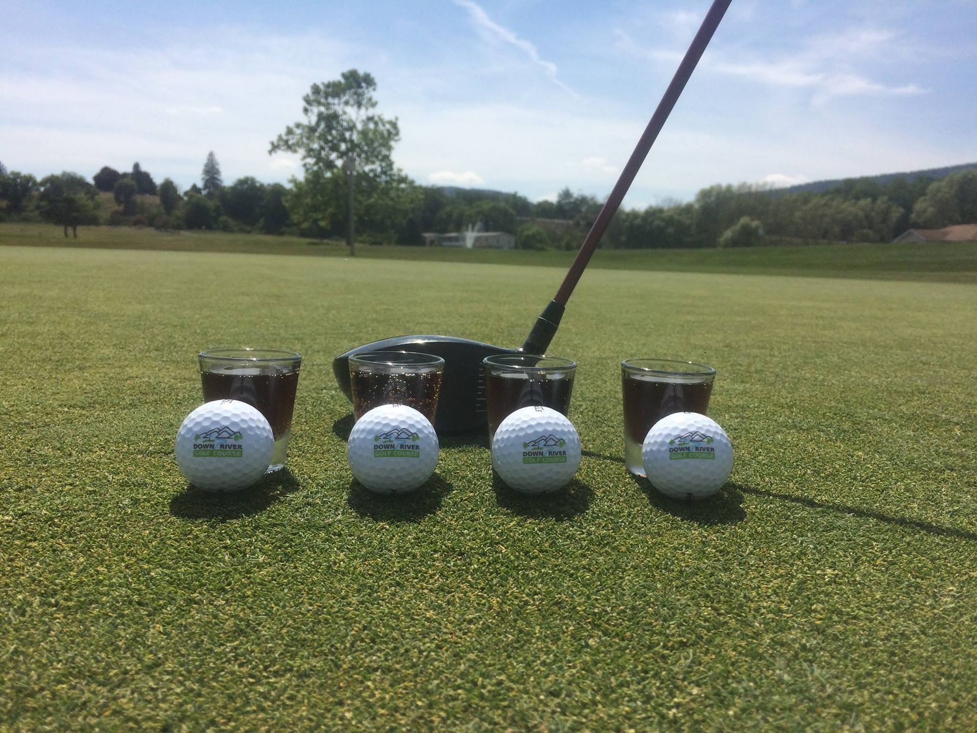 Four golf balls and shot glasses are on a green golf course with a golf club in the background.