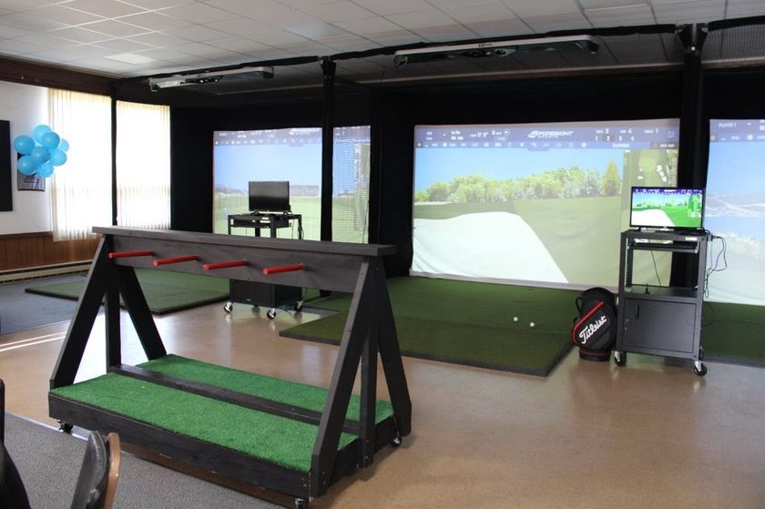 Indoor golf simulator room with three bays, screens displaying golf courses, and a golf club rack.