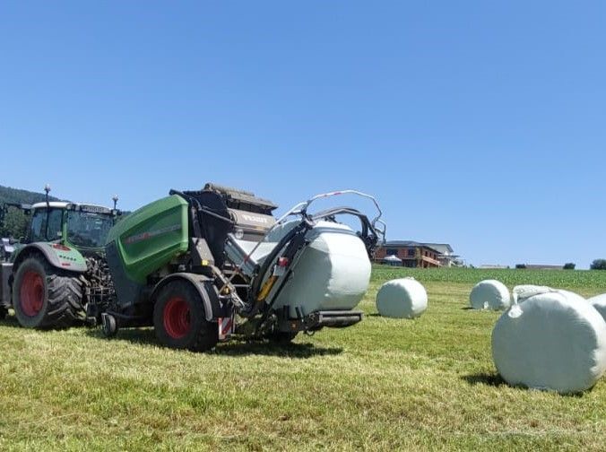Ein Traktor wickelt auf einem Feld einen Heuballen ein; im Hintergrund sind unter blauem Himmel mehrere eingewickelte Ballen zu sehen.